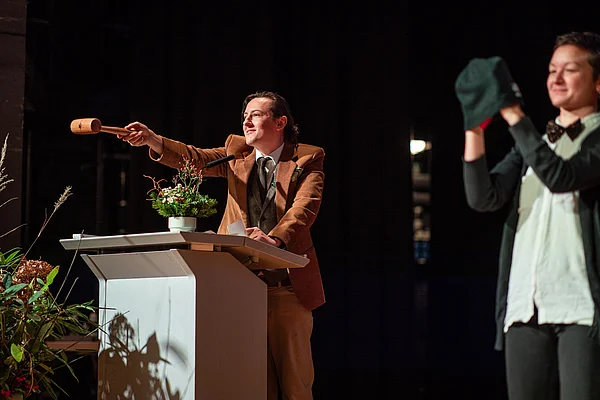 Eine Person hält einen Auktionshammer während eine zweite Person das Objekt (Mütze) präsentiert.