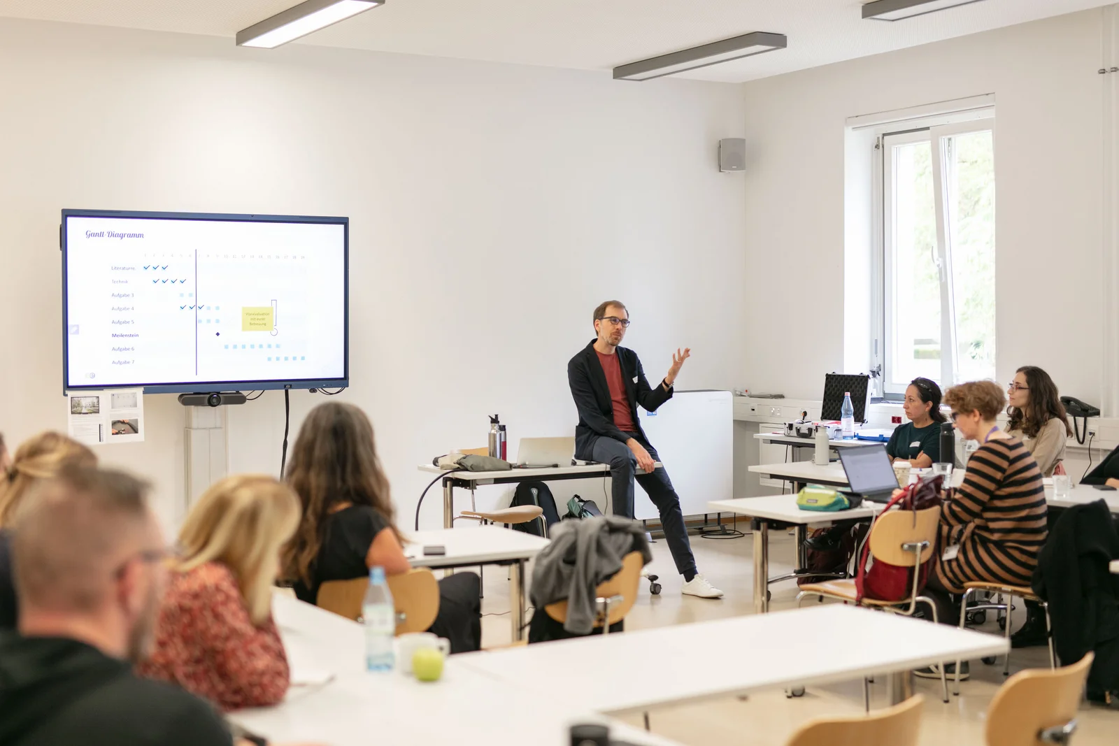 Ein Mann hält einen Vortrag, im Raum sitzen Zuhörer an Tischen.
