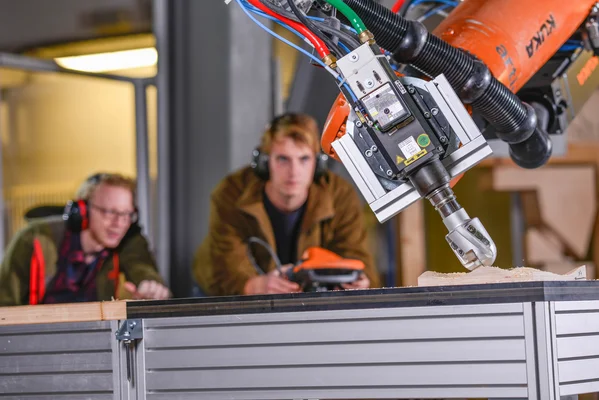 Roboterarm mit eingespanntem Fräskopf bearbeitet einen Holzprobekörper. Im Hintergrund sieht man zwei junge Wissenschaftler mit der tragbaren Robotersteuerung in der Hand.