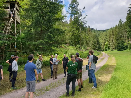ein Gruppe Studierender steht am Waldrand und hört einer Erklärung zu. Zwei Personen befinden sich im Hochsitz, der hier besprochen wird. 