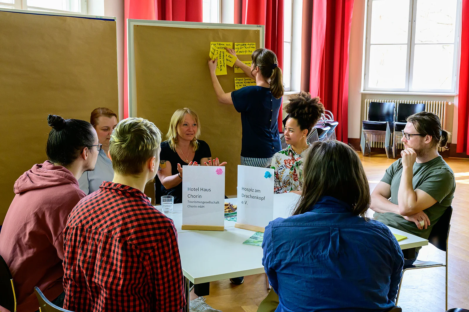 Studierende und zwei Unternehmensvertreterinnen tauschen sich an einem großen Tisch aus. Im Hintergrund werden Moderationskarten aufgehangen.