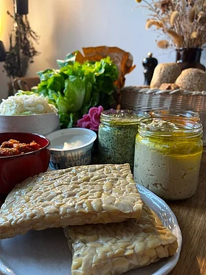 Tisch mit der Sammlung an Zutaten für das Tempeh-Brötchen, das im Rahmen des Moduls „Studienpartner Ökobranche“ (2.FS) entwickelt wurde. Man sieht den Tempeh, Salat, verschiedene Aufstriche, Sauerkraut und die Brötchen.
