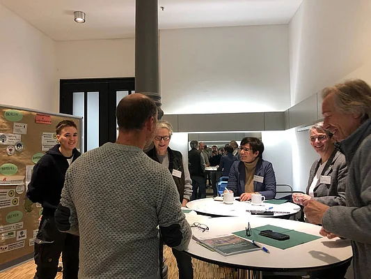 Sechs Personen stehen an einem Stehtisch und unterhalten sich.