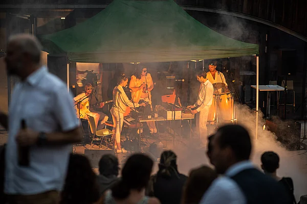 Publikum vor der fünfköpfigen Band Phase Odyssey auf der HNEE Abschlussfeier im Jahr 2022 unter einem Pavillon vor der Waldcampus Mensa, mit viel Elektronik, Live Instrumenten, weißer Kleidung, gehüllt in Rauch
