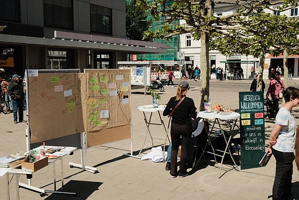 Set-Up einer Intervention auf dem Marktplatz im Rahmen des Moduls „Ernährungskulturen der Zukunft“ (4. FS). Man sieht das Begrüßungsschild, den Materialtisch und zentral den Traumbaum für die Visionselemente der BürgerInnen für die Ernährungskultur in Eberswalde 2040.