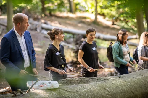 Fünf Personen sind im Wald und halten Drumsticks eines Schlagzeugs in den Händen, bereit auf einem Baumstamm zu trommeln.